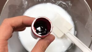 A Hand Holding an Open Bottle of Dark Red Food Coloring Above a Bowl of White Glue with A Spatula Inside